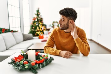 Sticker - Arab young man sitting on the table by christmas tree smiling with hand over ear listening an hearing to rumor or gossip. deafness concept.