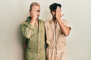 Wall Mural - Homosexual gay couple standing together wearing casual jumpsuit covering one eye with hand, confident smile on face and surprise emotion.