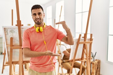 Wall Mural - Young hispanic man at art studio gesturing with hands showing big and large size sign, measure symbol. smiling looking at the camera. measuring concept.