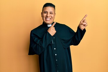 Young latin priest man standing over yellow background smiling and looking at the camera pointing with two hands and fingers to the side.