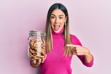 Young hispanic woman holding jar of chocolate chips cookies smiling happy pointing with hand and finger