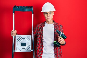 Poster - Young hispanic man holding screwdriver wearing hardhat by construction stairs in shock face, looking skeptical and sarcastic, surprised with open mouth