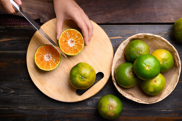 Hand holding knife and cutting green tangerine orange fruit on wooden board, Table top view