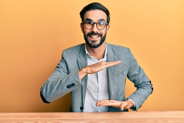 Wall Mural - Young hispanic man working at the office gesturing with hands showing big and large size sign, measure symbol. smiling looking at the camera. measuring concept.