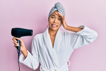 Poster - Young chinese woman wearing shower towel cap and bathrobe holding dryer stressed and frustrated with hand on head, surprised and angry face