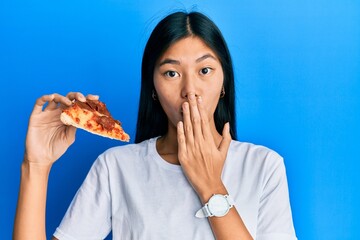 Wall Mural - Young chinese woman eating tasty pepperoni pizza covering mouth with hand, shocked and afraid for mistake. surprised expression