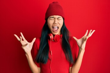 Poster - Young chinese woman listening to music using headphones crazy and mad shouting and yelling with aggressive expression and arms raised. frustration concept.