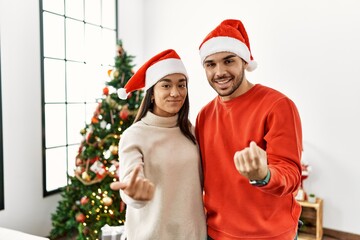 Poster - Young hispanic couple standing by christmas tree beckoning come here gesture with hand inviting welcoming happy and smiling