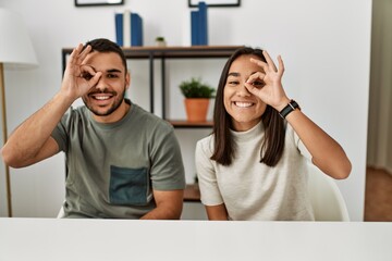 Sticker - Young latin couple wearing casual clothes sitting on the table smiling happy doing ok sign with hand on eye looking through fingers