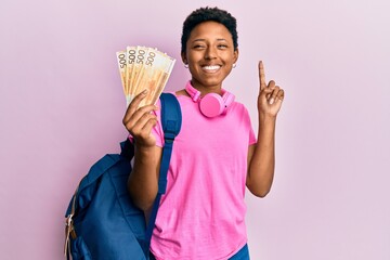 Wall Mural - Young african american girl wearing student backpack holding norwegian krone banknotes smiling with an idea or question pointing finger with happy face, number one