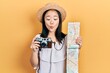 Young chinese girl holding city map and vintage camera making fish face with mouth and squinting eyes, crazy and comical.