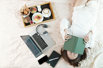 Wall Mural - Lazy young woman lying on bed with opened laptop and tray with breakfast, sleeping after reading boring book