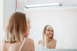 Young smiling woman apply skincare moisturizer body cream on face in bathroom after shower. Happy teenage girl do Self Care morning routine. Beauty spa treatment at home.