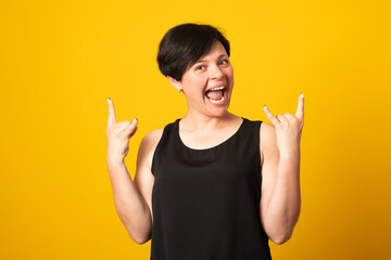 Emotive woman makes rock n roll sign, says I will rock this party, yells loudly, casual t shirt, stands over yellow background, feels self confident.