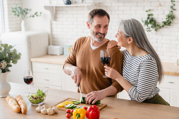 Relaxed mature middle-aged couple cooking together romantic dinner for date at home in the kitchen, drinking wine. Happy wife and husband preparing food. Vegetarian healthy eating