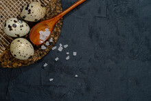 Three Quail Eggs And The Wooden Spoon Placed  On The Cork Supply. Slate Black Uneven Stone Background. Close Up. Top View. Free Space For Text Or Menu. Eco-Friendly Concept.