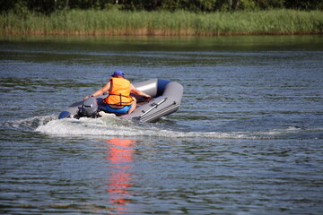 Wall Mural - One man in orange lifejacket turns on inflatable motor boat with small outboard motor on grassy far shore background , active recreation on the water at Sunnny summer day