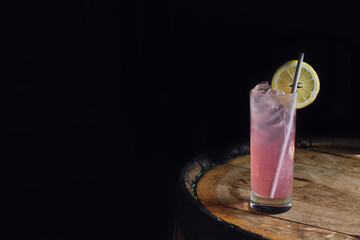 Pink lemonade in tall Collins glass with paper straw and lemon wheel on whiskey barrel against dark background