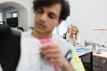 young dressmaker sitting near cloth sample and sewing machine while designer working on blurred foreground