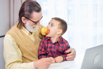 Handsome grandfather and grandson use gadgets and smile while spending time together at home. The grandson is playing with an apple