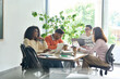 © insta_photos - Young happy creative team group four multicultural coworkers students brainstorming working on project together sitting in classroom office using laptops computers near panoramic window.