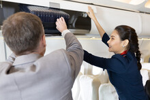 Flight Attendant Helping Passenger With Carry On Luggage To Overhead Bins
