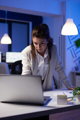 Focused tired businesswoman reading emails on laptop standing near desk in start-up company late at night. Focused employee using technology network wireless doing finance research in office