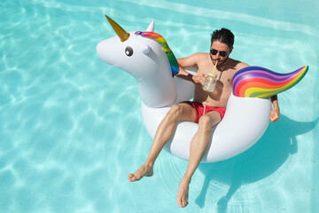 Top view of a young man wearing sunglasses drinking a refreshment on a unicorn inflatable ring in a swimming pool. Summer concept.