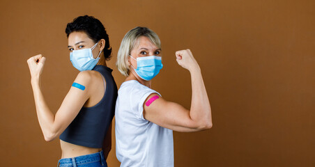 Wall Mural - Two gender female citizens wears face mask standing look at camera lean on each other back hold fist up showing strong muscle with colorful plaster together after vaccination