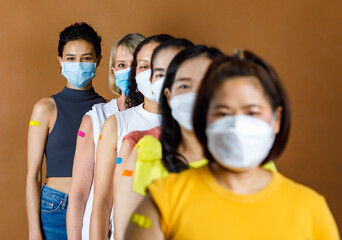 Diversity group of female from multinational ethnic patients wears face mask stand in line by height order look at camera showing colorful plasters on shoulder together after vaccination