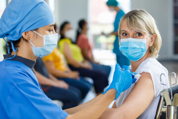 Wall Mural - Asian female doctor wears face mask and blue hospital uniform injecting vaccine to Caucasian senior woman at ward working desk with equipment while other patients wait in queue in blurred background