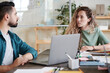 © AnnaStills - Young businesswoman sitting at the table and talking to her colleague they using laptop in their work at office