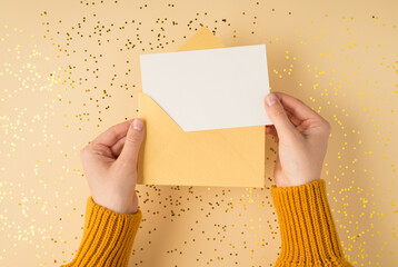 First person top view photo of female hands in yellow pullover holding open pastel yellow envelope with white card over scattered golden sequins on isolated pastel orange background with blank space