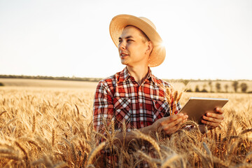 Wall Mural - Portrait of a young farmer in a straw hat stands with a tablet and spikelets in his hands in the middle of a ripe wheat field. Caucasian man checking quality and growth of crops for agriculture.