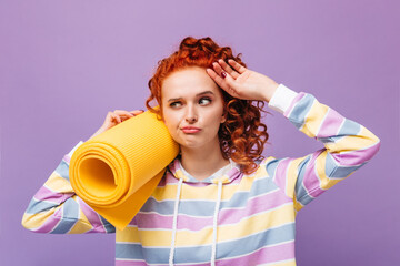 Wall Mural - Red-haired girl in multi-colored hoodie touches her forehead and holds yellow mat for sports