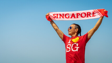 Singapore Day. Joyful middle aged man holds scarf with the words Singapore over his head. Asian handsome male smiles on blue sky background. Celebration of Independence day. National event. Festival.