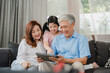 © tirachard - Asian grandparents and granddaughter using tablet at home. Senior Chinese, grandpa and grandma happy spend family time relax with young girl checking social media, lying on sofa in living room concept