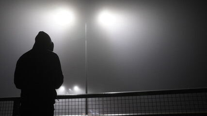 A mysterious, lonely hooded figure, back to camera. Standing on a bridge, over looking a motorway on a misty winters at night.