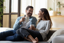 Calm Relaxed Couple Hold Cups Rest On Comfy Sofa Enjoy Carefree Talk While Sit Together On Couch At Modern Living Room. Homeowners Family Spend Weekend At Home, Morning Beverage, Communication Concept