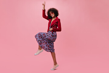 Poster - Active girl in red jacket and flowered dress shows peace sign on pink background. Cool curly woman in floral skirt jumping on isolated backdrop