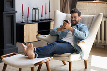 Handsome 30s Single Man Relaxing On Comfy Armchair Put Feet On Footstool Holding In Hands Smartphone Spend Weekend Alone In Cozy Cottage House. No Stress, Home Owner, Modern Tech Usage, Fun Concept