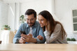 © fizkes - Mixed race couple sit in kitchen at table looking at smartphone screen interested with good e-commerce offer, sale low price and discount in retail services, buying ticket. New mobile app, fun concept