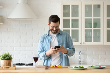 Handsome millennial single man cooking in modern kitchen holds smart phone searching recipes on internet, looking at device screen smile make food products delivery order, enjoy easy on-line services