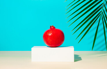 Wall Mural - Minimal summer composition with  pomegranate fruit standing on a white pedestal and a palm branch against a blue background. Creative tropical or food concept with copy space.