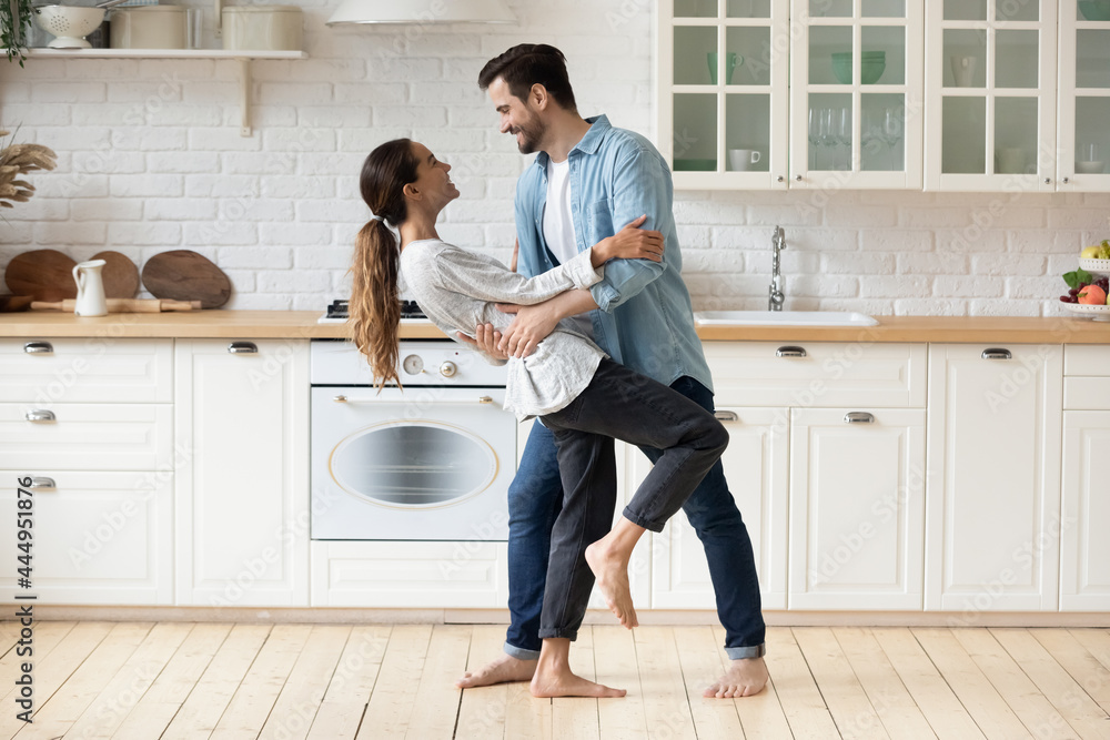 Young beautiful mixed race couple home owners family celebrate relocation day housewarming dancing in modern cozy kitchen standing barefoot on warm floor feel happy. Romance, dating, bank loan concept - obrazy, fototapety, plakaty 