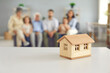 © Studio Romantic - Closeup of small little toy house on table against blurred background of living-room with whole big family of mom, dad, grandma, grandpa and kids sitting together. Real estate, buying new home concept