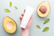 Natural Avocado Skincare Products For Healthy Skin And Hair. Woman Hand Holding Blank White Squeeze Tube With Fresh Organic Avocado On Green Background. Beauty Herbal Skincare And Haircare Concept.