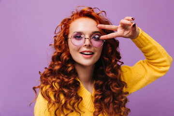 Wall Mural - Charming woman in round glasses and yellow sweater poses on lilac background and shows peace sign