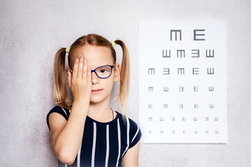 Wall Mural - Little girl wearing eyeglasses taking eyesight test before school with blurry eye chart at the background