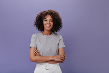 Wall Mural - Portrait of young girl in gray t-shirt on purple background. Closeup snapshot of curly brunette woman in light outfit smiling on isolated backdrop